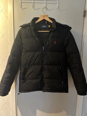 Svart pufferjacka från Polo Ralph Lauren - Snygg svart pufferjacka från Polo Ralph Lauren med huva och dragkedja framtill. Jackan har två framfickor med dragkedja och den klassiska röda Polo-loggan på bröstet. Perfekt för kalla vinterdagar och riktigt skön att bära.