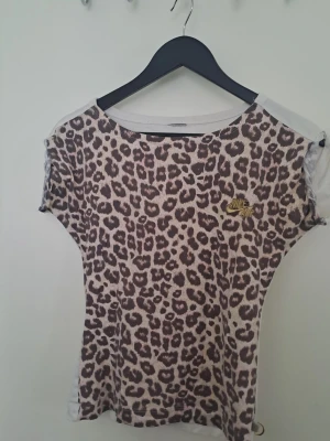 Nike t-shirt med leopardmönster - T-shirt från Nike med leopardmönstrad framsida och vit baksida. T-shirten har korta ärmar och guldig Nike-logga på bröstet. Se bilder för skick. 🩷 Uppskattad storlek xs-s