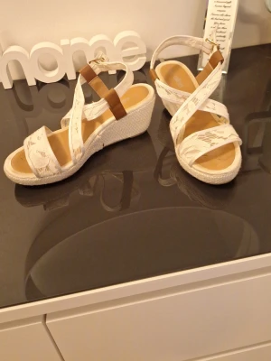 Vita kilklackade sandaler med spets strl 37 - Säljer ett par vita sandaler med kilklack och snygga spetsdetaljer. Remmarna är i vitt tyg med spetsmönster och en brun detalj vid ankeln. Klacken är flätad i repstil för en fräsch sommarkänsla. Perfekta för dig som vill ha både höjd och komfort. Använts en hel del vid skolavslutningar 