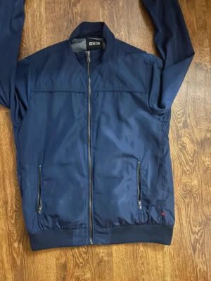 Mörkblå bomberjacka Big Star - Snygg mörkblå bomberjacka från Big Star med dragkedja framtill och två fickor med dragkedja. Jackan har ribbade muddar vid ärmslut och nederkant samt en klassisk bomberkrage. Materialet är slätt och lätt, perfekt för en cool streetstil.