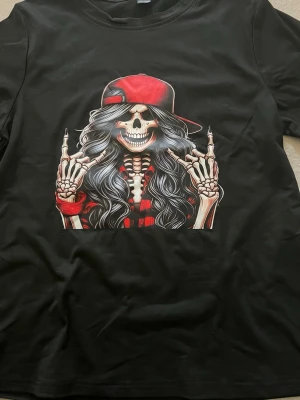 Svart t-shirt med skelettmotiv storlek XL - Cool svart t-shirt med tryck av ett skelett med långt hår, röd keps och röd skjorta. Motivet är detaljerat och sticker ut med sina kontrasterande färger. Perfekt för dig som gillar streetwear och unika prints.