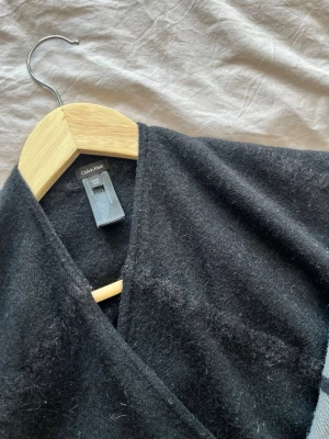 Svart och grå halsduk Calvin Klein - Poncho från Calvin Klein i svart och grått med rutmönster på ena sidan och fransar i ändarna 