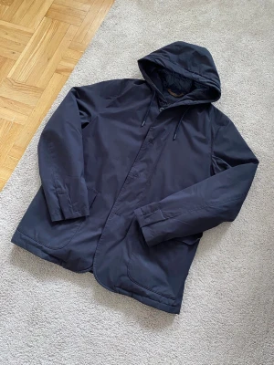 Massimo Dutti Kavaj Dunjacka - Stilren svart Dunjacka/ tjockare Skjortjacja med huva och dragsko. Jackan har dragkedja och knappar framtill samt två sidofickor. Materialet är slätt och vattenavvisande, perfekt för kyliga dagar. Passar dig som vill ha en enkel och funktionell jacka med modern look.