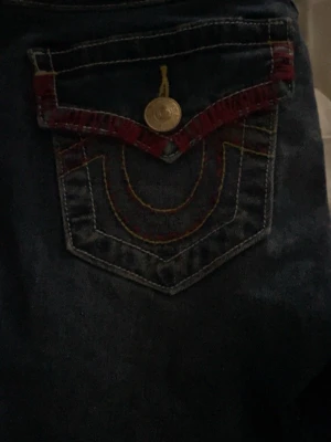 True Religion Joey Low Rise Flare Jeans - Säljer ett par mörkblå True Religion Joey jeans med låg midja och utsvängda ben. Snygga detaljer med guldfärgad knapp, röd och gul kontrastsöm samt dekorativ ficka bak. Klassisk True Religion-stil som sticker ut.