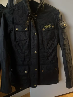 Svart jacka från Barbour - Snygg svart jacka från Barbour International med gulddetaljer och flera fickor. Jackan har hög krage, tryckknappar och ett klassiskt Barbour-märke på bröstet. 