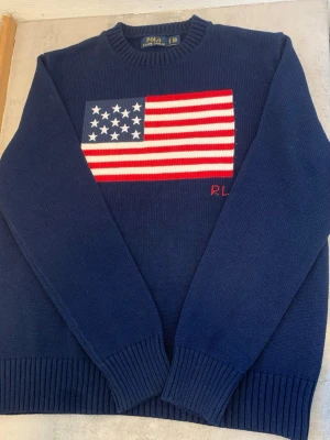 Blå stickad tröja Polo Ralph Lauren - Mörkblå stickad tröja från Polo Ralph Lauren med amerikansk flagga i rött, vitt och blått på bröstet. Rund halsringning och ribbade muddar vid ärmslut och nederkant. Klassisk och stilren design som sticker ut. Storlek M. Är i rejält bra skick.