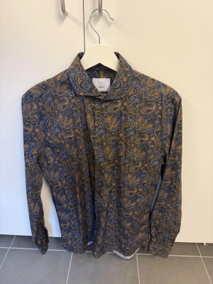 Mönstrad skjorta från Bläck, stl S - Snygg långärmad skjorta från Bläck med blå bas och paisley-inspirerat mönster i brunt och gult. Skjortan har klassisk krage och knäppning framtill. Tillverkad i mjuk bomull som känns skön mot huden. Perfekt för dig som vill sticka ut med en unik stil.