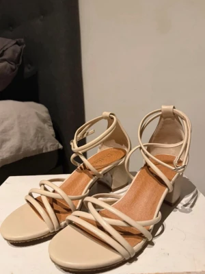 Beige sandaletter med blockklack - Säljer ett par stilrena beige sandaletter med tunna remmar och blockklack. Skorna har öppen tå och justerbart spänne runt ankeln. Tillverkade i syntetmaterial med en mjuk innersula för extra komfort. Perfekta för dig som gillar minimalistisk och trendig stil. Använda endast engång 