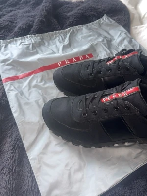 Svarta Prada sneakers  - Snygga svarta sneakers från Prada med klassisk låg siluett. Skorna har en röd och vit Prada-logga på plösen, svarta snören och detaljer i textil och mocka. Perfekta för dig som vill ha en clean och exklusiv look.