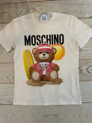 Vit Moschino t-shirt med björntryck - Säljer en vit t-shirt från Moschino med ett stort tryck av en nallebjörn i semesteroutfit, surfbräda och sol i bakgrunden. T-shirten har rund hals och korta ärmar. Perfekt för dig som gillar lekfulla designer och vill sticka ut. Måtten är 47 cm axel till axel, längd 71 cm