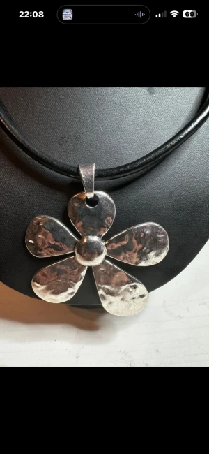 Stort halsband med silverfärgad blomma - Unikt halsband med en stor, hamrad blomma i silverfärgad metall som hänge. Blomman har fem breda kronblad och sitter på ett svart, rundat band i läder. Perfekt statement piece för dig som vill sticka ut med din stil.