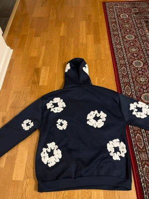 Mörkblå denim tears hoodie - Snygg mörkblå hoodie med vita blommönster utspridda över hela tröjan, även på huvan. Hoodien har en klassisk känguruficka framtill och ribbade muddar vid ärmslut och nederkant. Perfekt för dig som vill sticka ut med en unik och trendig stil.
