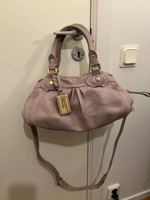 Ljuslila handväska från Marc by Marc Jacobs - Säljer en ljuslila handväska från Marc by Marc Jacobs i mjukt, strukturerat läder. Väskan har både handtag och justerbar axelrem samt guldfärgade metalldetaljer och en ikonisk metallbricka framtill. Perfekt för dig som vill ha en stilren och trendig accessoar.