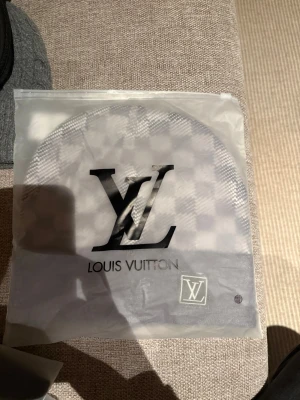 Grå Louis Vuitton mössa - Snygg grå mössa från Louis Vuitton med klassiskt rutmönster och vikt kant. Mössan har en diskret LV-logga framtill och är tillverkad i mjukt material som känns skönt mot huden. Perfekt accessoar för dig som gillar exklusiv streetstyle.