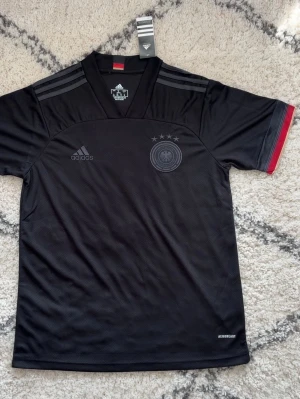 Svart Tyskland matchtröja Adidas - Snygg svart matchtröja från Adidas med Tysklands landslagslogga och tre stjärnor på bröstet. Tröjan har V-ringning, korta ärmar och diskreta grå ränder på axlarna. Röd och gul detalj på ärmslutet och tillverkad i lätt, ventilerande material (AEROREADY).