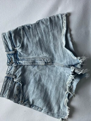 Ljusblå denimshorts med fransar - Säljer ett par ljusblå denimshorts med hög midja och råa fransade benslut. Klassisk femficksmodell med dragkedja och knapp framtill. Perfekta för sommaren och ger en avslappnad look.