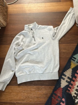 Grå halvzip från Ralph Lauren - Snygg ljusgrå halvzip-tröja från Ralph Lauren med liten broderad logga på bröstet. Tröjan har hög krage, dragkedja upptill och ribbade muddar vid ärmslut och midja. Perfekt till jeans eller chinos för en clean look.