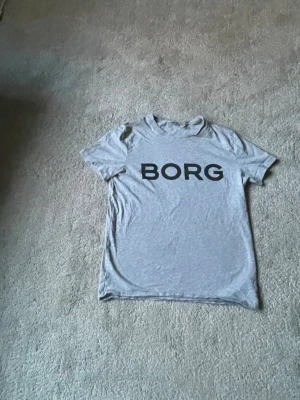 Grå t-shirt från BORG - Snygg grå t-shirt från BORG med stor svart logga över bröstet. Klassisk rund halsringning och korta ärmar. Tillverkad i mjukt bomullsmaterial som känns skönt mot huden. Perfekt för en avslappnad och stilren look.40kr frakt bitalas av köpare.