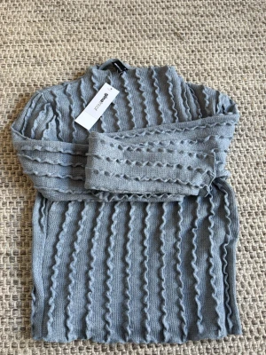 Ljusblå stickad tröja från Kompagniet - Trendig ljusblå stickad tröja från Kompagniet med vågigt strukturmönster över hela plagget. Tröjan har lång ärm och hög hals, perfekt för lager på lager. Materialet känns mjukt och bekvämt, och färgen är lätt att matcha till olika outfits.