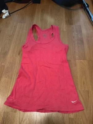 Rosa Nike Dri-FIT träningslinne - Snyggt rosa träningslinne från Nike med Dri-FIT-material som andas. Linnet har brottarrygg och en liten vit Nike-logga framtill. Perfekt för gymmet eller löprundan när du vill ha något bekvämt och sportigt.