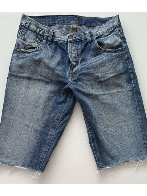 Blå jeansshorts från GUL & BLÅ, W31 - Tidlösa blå stentvättade jeansshorts från anrika Gul & Blå med knappgylf, storlek W31 (S/M). Shortsen har en klassisk design med fem fickor, inklusive en myntficka framtill. Bakfickorna har dekorativa sömmar och en liten logotypetikett. Midjebandet har hällor för bälte och en stor logotyp i läderimitation baktill. Mycket gott skick, som nya!