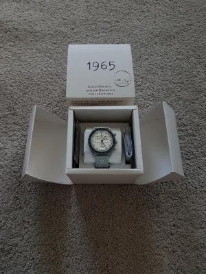 Omega x Swatch MoonSwatch 1965 - Säljer en Omega x Swatch Mission to The Moon 1965 MoonSwatch från Bioceramic-kollektionen. Klockan har en ljus urtavla med svarta detaljer och grått armband. Klassisk kronografdesign med tachymeter och Omega-logga på armbandet. Perfekt för dig som gillar stilrena accessoarer.