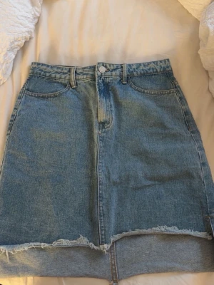 Blå jeanskjol med rå kant - Säljer en blå jeanskjol i klassisk modell med fem fickor och bälteshällor. Kjolen har en rå, fransig kant nedtill och en liten slits framtill för extra edge. Perfekt till vardags för en avslappnad look.