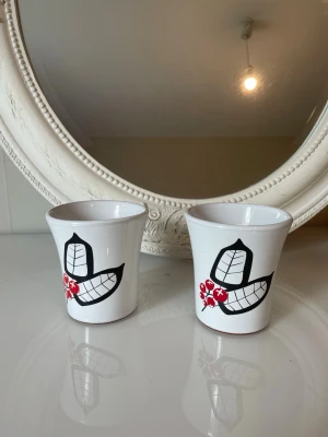 Två vita keramikmuggar med bärmotiv från rotor  - Säljer två vita keramikmuggar från rotor design med handmålat motiv av svarta blad och röda bär. Muggarna har en klassisk form och är glaserade med en matt terrakottafärgad kant i botten. Perfekta för kaffe eller te och ger en retro känsla till köket.