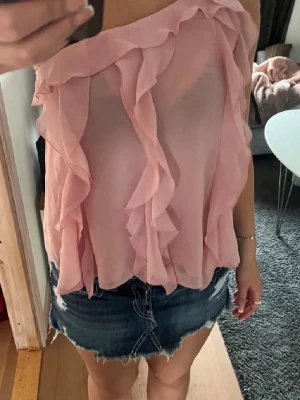 Rosa Zara volanglinne  - Säljer en rosa, axelbandslös tubtopp med volangdetaljer. Toppen är gjord i ett lätt och transparent material som ger en luftig känsla. Perfekt för dig som gillar att sticka ut med en feminin och trendig look.