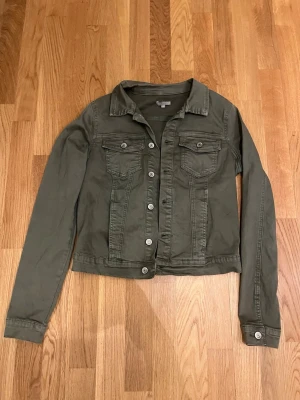 Olivgrön jeansjacka med fickor - Snygg olivgrön jeansjacka med klassisk krage och silverfärgade knappar. Två bröstfickor med lock och knapp, samt långa ärmar med knapp vid ärmslut. Stretchig och följsam passform och stilren design som passar till det mesta. Bröst omkrets 94cm. Fint skick med en sliten look. 