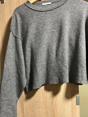 Grå croppad stickad tröja Zara - Snygg gråmelerad croppad stickad tröja från Zara med rund halsringning och avslappnad passform. Tröjan har långa ärmar och mjukt material som passar perfekt till jeans eller kjol. Enkel och stilren design för en trendig look. 