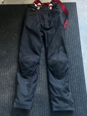 Svarta täckbyxor med hängslen - Säljer ett par svarta byxor med knäskydd för dig som älskar åka eller köra motorcykel. Från Didriksons med röda justerbara hängslen och förstärkta knän. Byxorna har dragkedja fram, ficka på sidan och slitstarka detaljer. Perfekta för kalla dagar och vinteraktiviteter.