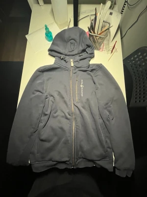 Mörkblå hoodie från Sail Racing - Mörkblå hoodie från Sail Racing med dragkedja framtill och en bröstficka med dragkedja. Märkets logga är tryckt på bröstet. Hoodien har huva och ribbade muddar vid ärmslut och nederkant. Tillverkad i mjukt bomullsmaterial för skön känsla.