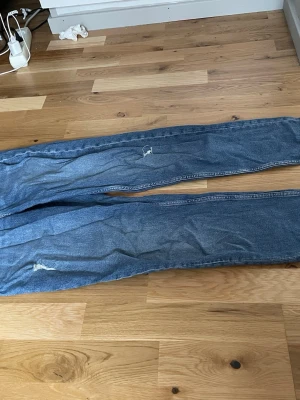 Blå raka jeans med slitningar - Säljer ett par klassiska blå jeans med raka ben och slitningar på både lår och knä. Jeansen har en avslappnad passform och är tillverkade i denim. Perfekta för en casual och trendig look.