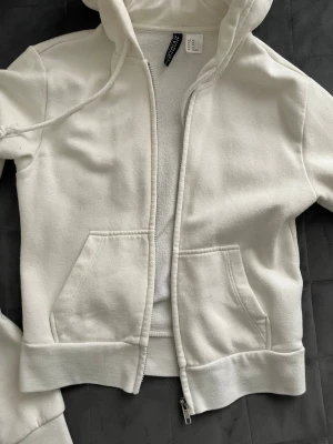 Vit kofta - Klassisk vit kofta med dragkedja framtill, två  fickor och justerbar huva med snören. Tillverkad i mjukt material som känns skönt mot huden. Perfekt för en avslappnad och sportig stil.