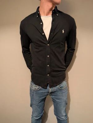 Svart skjorta från Ralph Lauren - Klassisk svart skjorta från Ralph Lauren med knappslå och ikoniska broderade loggan på bröstet. Skjortan har lång ärm, normal passform och är tillverkad i mjuk bomull. Perfekt till jeans eller chinos för en clean look.