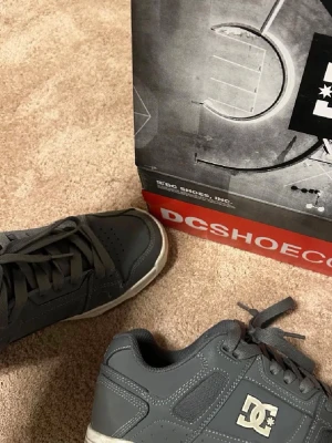 Mörkgrå DC Shoes sneakers - Säljer ett par mörkgrå DC Shoes sneakers med vita detaljer och logga på sidan och hälen. Skorna har klassisk låg profil, tjock vadderad plös och krage samt slitstark gummisula. Perfekta för dig som gillar streetwear och skate-inspirerad stil.