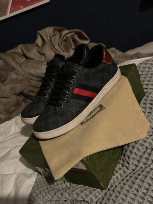 Gucci sneakers med monogram och röd detalj - Snygga Gucci sneakers i mörkgrått med klassiskt GG-monogram, svarta skosnören och vit sula. Sidan har en röd och marinblå rand, samt en metallicröd detalj på hälen. Perfekta för dig som vill ha en lyxig och trendig look.kommer med endast dustbag