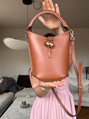 Brun axelväska - Snygg brun bucket axelväska med justerbar axelrem och guldfärgat spänne framtill. Väskan har en stilren, rundad form och kontrastsömmar som ger en modern touch. Perfekt för dig som vill ha en trendig och praktisk väska till vardagslooken💓💓 den är ny och har inga skador!!💕✌🏽✌🏽🌟