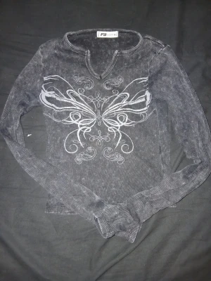 Svart långärmad topp med fjäril - Cool svart långärmad topp från FB Sister i ribbad bomull med vit fjärilsprint framtill. Toppen har en lätt tvättad look och v-ringad hals. Perfekt för dig som gillar unika detaljer och vill sticka ut lite extra.