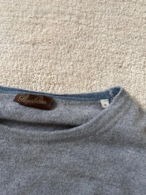 Stenströms merino tröja - Säljer en stilren grå stickad tröja från Stenströms med rund halsringning och bruna mockadetaljer på armbågarna. Perfekt för dig som gillar klassisk och enkel design. Tröjan har ribbade muddar vid ärmslut och nederkant.