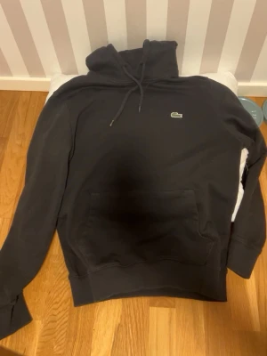 Svart hoodie från Lacoste - Säljer en svart hoodie från Lacoste med klassisk krokodillogga på bröstet. Hoodien har huva med dragsko och en stor magficka framtill. Perfekt för dig som gillar enkel och stilren streetwear.