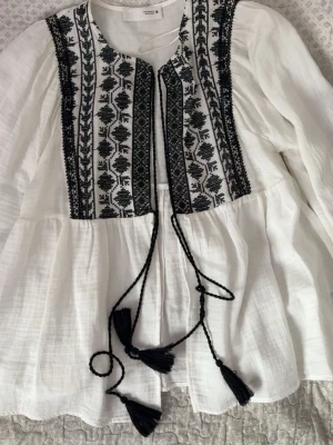 Vit boho-blus med brodyr från H&M - Supersnygg vit boho-inspirerad blus från ZARA med svart broderi framtill och dekorativa knytband med tofsar. Blusen har en lös passform, rund halsringning och långa ärmar. Tillverkad i ett tunt, luftigt material som känns skönt mot huden.