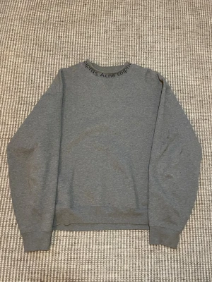 Grå sweatshirt från Acne Studios - Säljer en stilren grå sweatshirt från Acne Studios i 100% bomull. Tröjan har rund halsringning med Acne Studios-logga broderad runt kragen och ribbade muddar vid ärmslut och nederkant. Perfekt för en clean och avslappnad look.