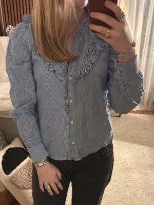 Blå- och vitrandig blus med volang från Holly & Whyte - Säljer en blå- och vitrandig blus från Holly & Whyte med volangdetaljer längs kragen och knäppning framtill. Blusen har långa ärmar och klassisk krage, perfekt för en stilren look. Nypris 400kr