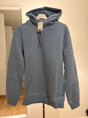Ljusblå hoodie med huva - Snygg ljusblå hoodie från H&M med klassisk känguruficka och dragsko i huvan. Hoodien har en avslappnad passform och ribbade muddar vid ärmar och nederkant. Perfekt för en chill och stilren look.