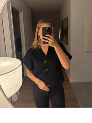 Svart topp Zara  - Stilren svart blus med v-ringning och två dekorativa knappar framtill från Zara. Modellen har korta och vida armar. Perfekt att matcha med både jeans och kjol. Säljer då den inte kommer till användning🤗
