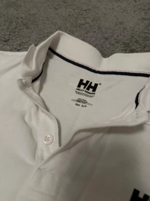Vit pikétopp från Helly Hansen - Klassisk vit pikétopp från Helly Hansen med svart broderad logga på bröstet. Topp med krage, knappar framtill och diskreta svarta detaljer längs insidan av kragen. Perfekt för en clean och sportig look.