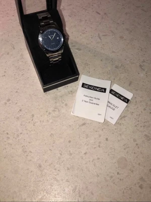 Sekonda silverfärgad klocka - Stilren klocka från Sekonda med silverfärgad länk i metall och mörkblå urtavla med diskreta ränder. Klockan har klassisk design och levereras i originalask med instruktionsbok. Perfekt accessoar för dig som gillar en clean look.