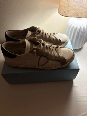 Beige sneakers med mörk blå detaljer - Snygga beige sneakers från Philippe Model med mörk blå hälparti och klassisk logotyp på sidan. Skorna har vita snören och är tillverkade i en mix av läder och textil. Perfekta för dig som gillar stilrena och trendiga sneakers.